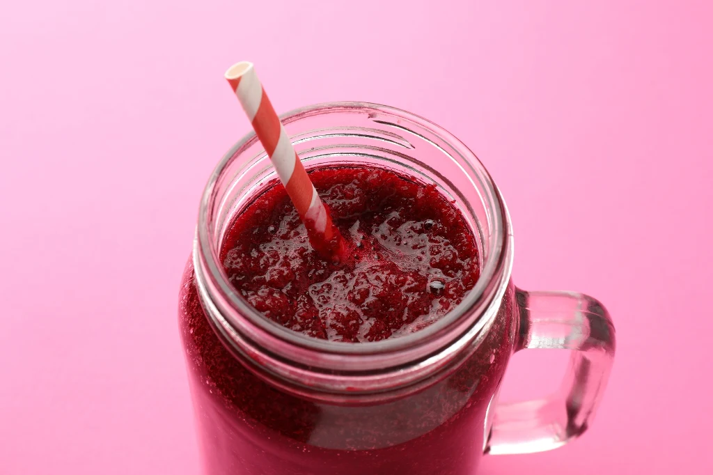 Szklany słoik wypełniony czerwonym smoothie lub koktajlem, przykryty papierową słomką w biało-czerwone paski, na jednolitym różowym tle.