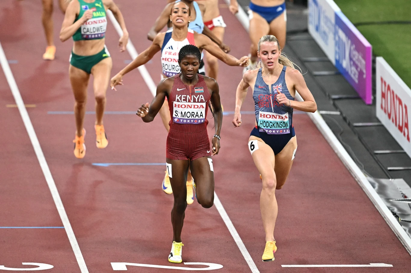 Zawodniczki biegnące w wyścigu na lekkoatletycznym stadionie, skupione na rywalizacji, w tle inni sportowcy, na ich strojach widoczne nazwiska oraz nazwy państw, wyraźnie widoczne linie torów.