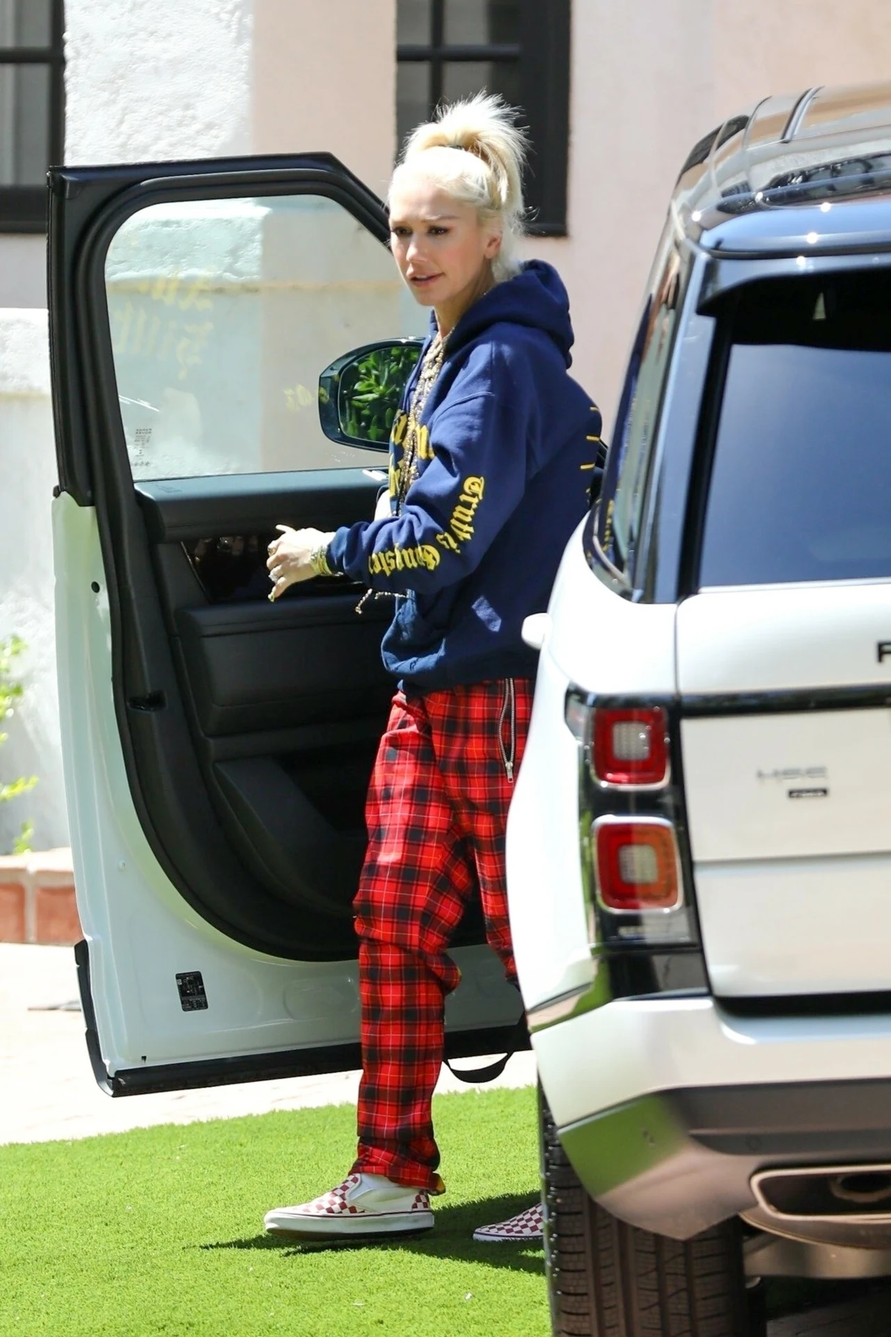 Gwen Stefani od lat jest wierna spodniom w kratę Kobieta o jasnych włosach ubrana w niebieską bluzę z kapturem oraz czerwone w kratę spodnie stoi obok otwartych drzwi białego samochodu terenowego na parkingu przy budynku.
