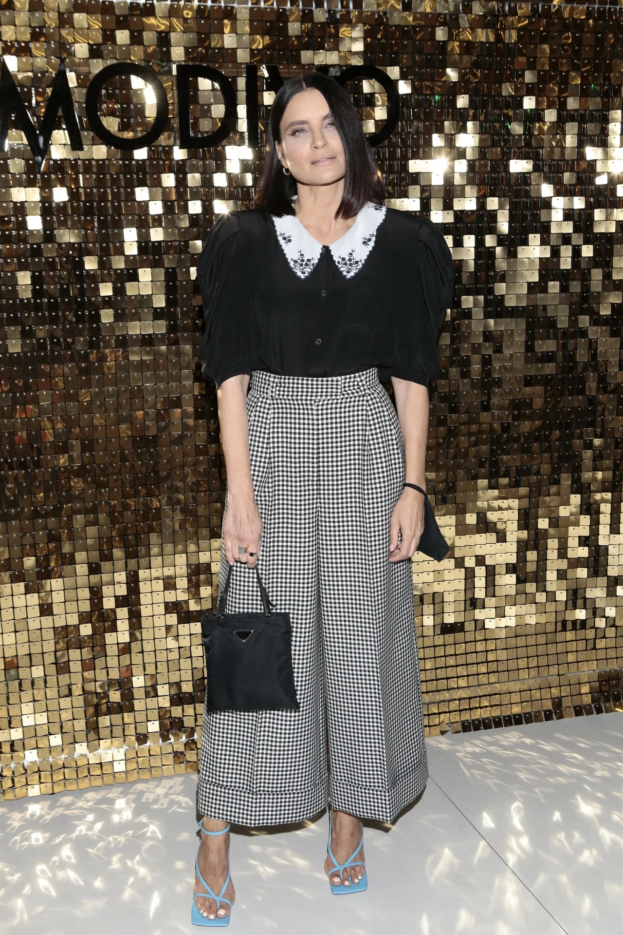 Joanna Horodyńska postawiła na kratę w eleganckiej odsłonie Kobieta o ciemnych, prostych włosach ubrana w czarno-białą stylizację z szerokimi spodniami w kratę, czarną bluzką z białym kołnierzykiem i jasnoniebieskimi sandałami na obcasie, trzyma czarną torebkę, stoi na tle połyskującej złotej ścianki dekoracyjnej.