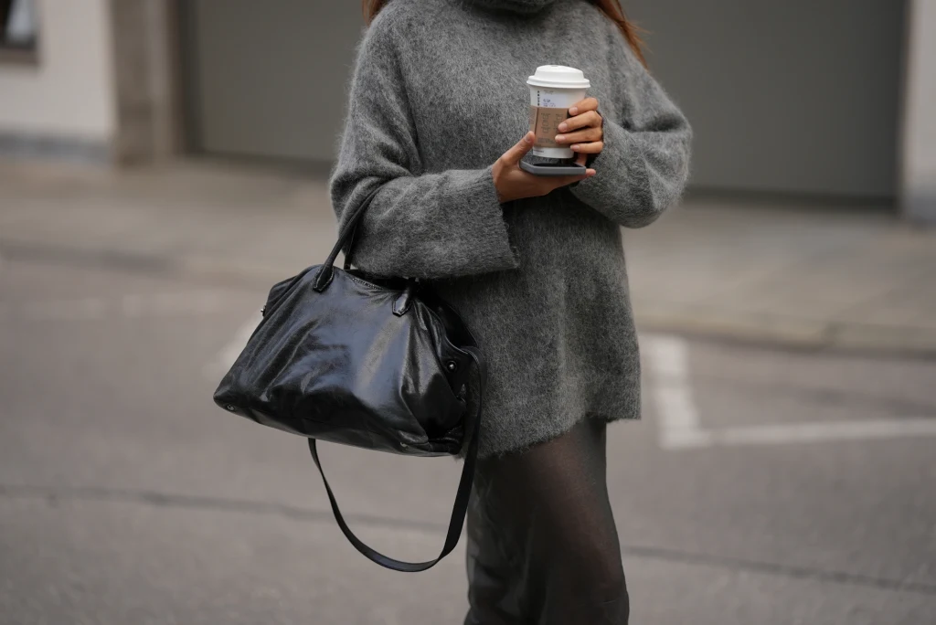 Kobieta ubrana w szary, oversize'owy sweter z kubkiem kawy w dłoni oraz dużą czarną torbą na ramieniu spaceruje ulicą.