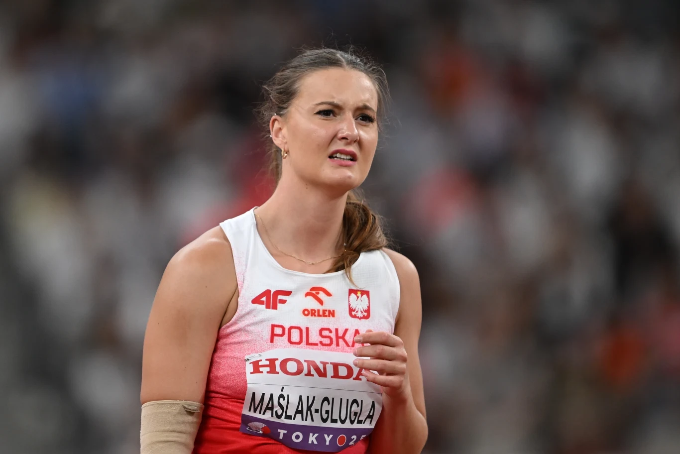 Małgorzata Maślak-Glugla Zawodniczka w stroju reprezentacji Polski podczas zawodów lekkoatletycznych na stadionie, widoczna tabliczka z nazwiskiem oraz logotypy sponsorów.