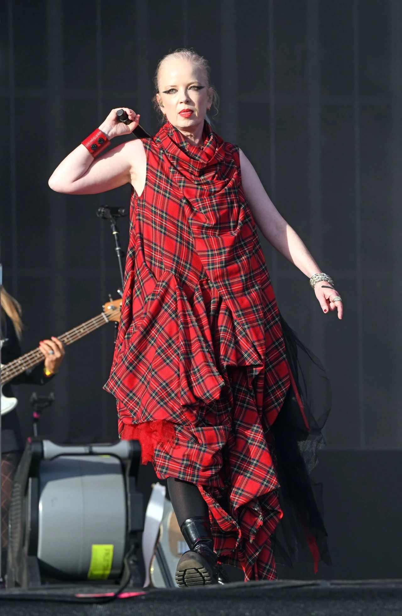 Shirley Manson kocha kratę w wersji total look Kobieta w ekstrawaganckiej, czerwonej sukni w szkocką kratę wykonuje występ na scenie muzycznej, trzymając mikrofon ciekawe, odważne stylizacje, wyrazista mimika twarzy, oświetlenie sceniczne.