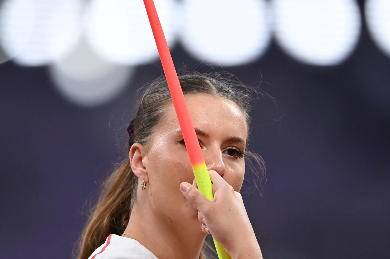 Małgorzata Maślak-Glugla Kobieta skupiona na rzucie oszczepem, trzymająca oszczep przed swoją twarzą na tle rozmazanego stadionu.