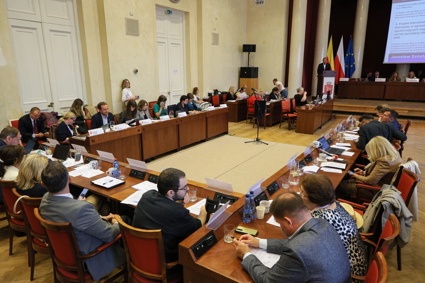 Sala obrad zasiadana przez uczestników przy dużych stołach ustawionych w podkowę, na stole dokumenty i mikrofony, z tyłu widać pulpit prowadzącego oraz flagi polską, miejską, unijną, uczestnicy skupieni na dyskusji.
