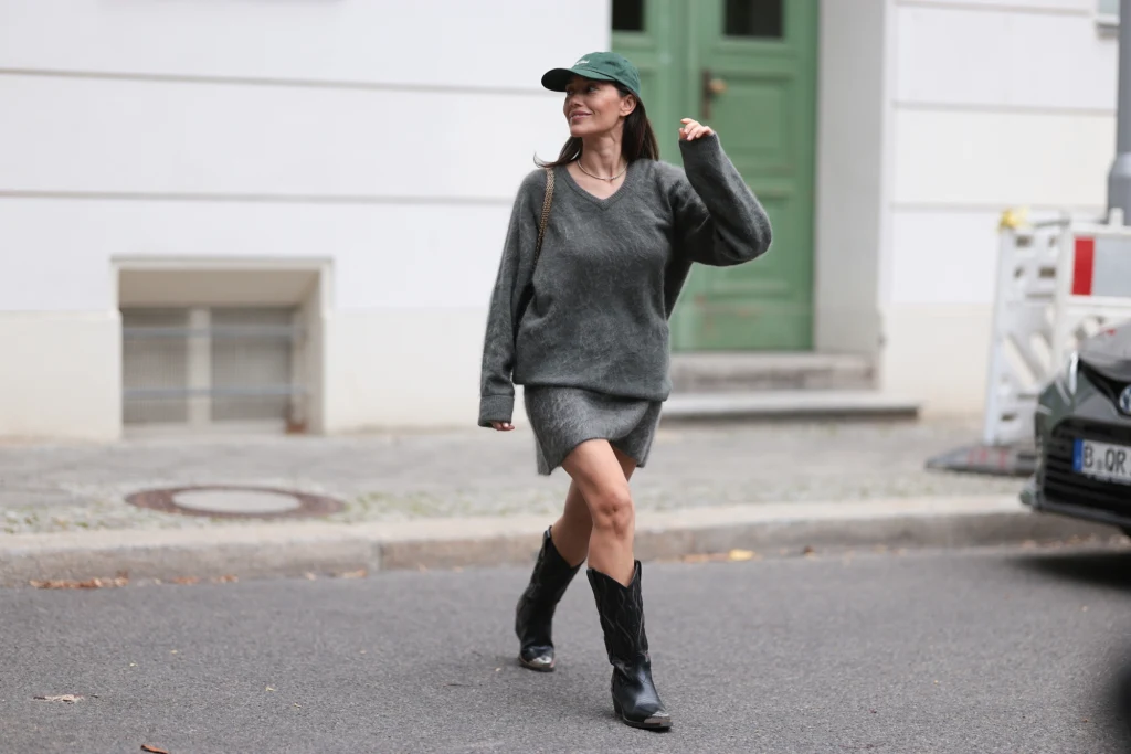Młoda kobieta ubrana w luźny ciemnoszary sweter oraz spódnicę mini w tym samym kolorze, na głowie zielona czapka z daszkiem, na nogach czarne kowbojki, idzie ulicą miasta przy białym budynku z zielonymi drzwiami.