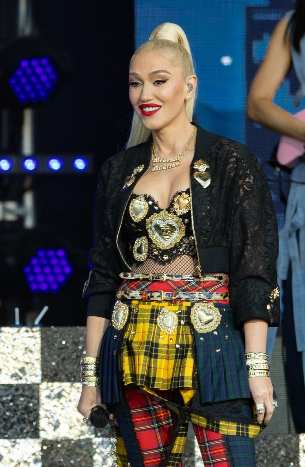 Gwen Stefani od lat stawia na kratę Kobieta z długimi blond włosami spiętymi w koński ogon uśmiecha się na scenie, ubrana w oryginalny, kolorowy strój sceniczny z elementami biżuterii przypominającej serca oraz wzorzystą spódnicę, trzyma mikrofon; w tle scena z oświetleniem.