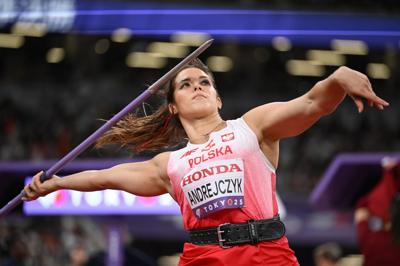 Maria Andrejczyk, Tokio 2025 Zawodniczka w stroju reprezentacji Polski wykonuje rzut oszczepem na stadionie lekkoatletycznym, w tle widoczne rozmyte światła i trybuny.