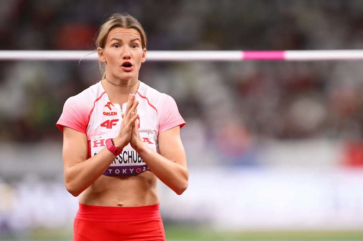 Lekkoatletka w czerwonym stroju sportowym stoi przed poprzeczką do skoku wzwyż, trzymając dłonie złączone jak do modlitwy, z wyrazem skupienia i emocji na twarzy, w tle rozmyte trybuny stadionu.