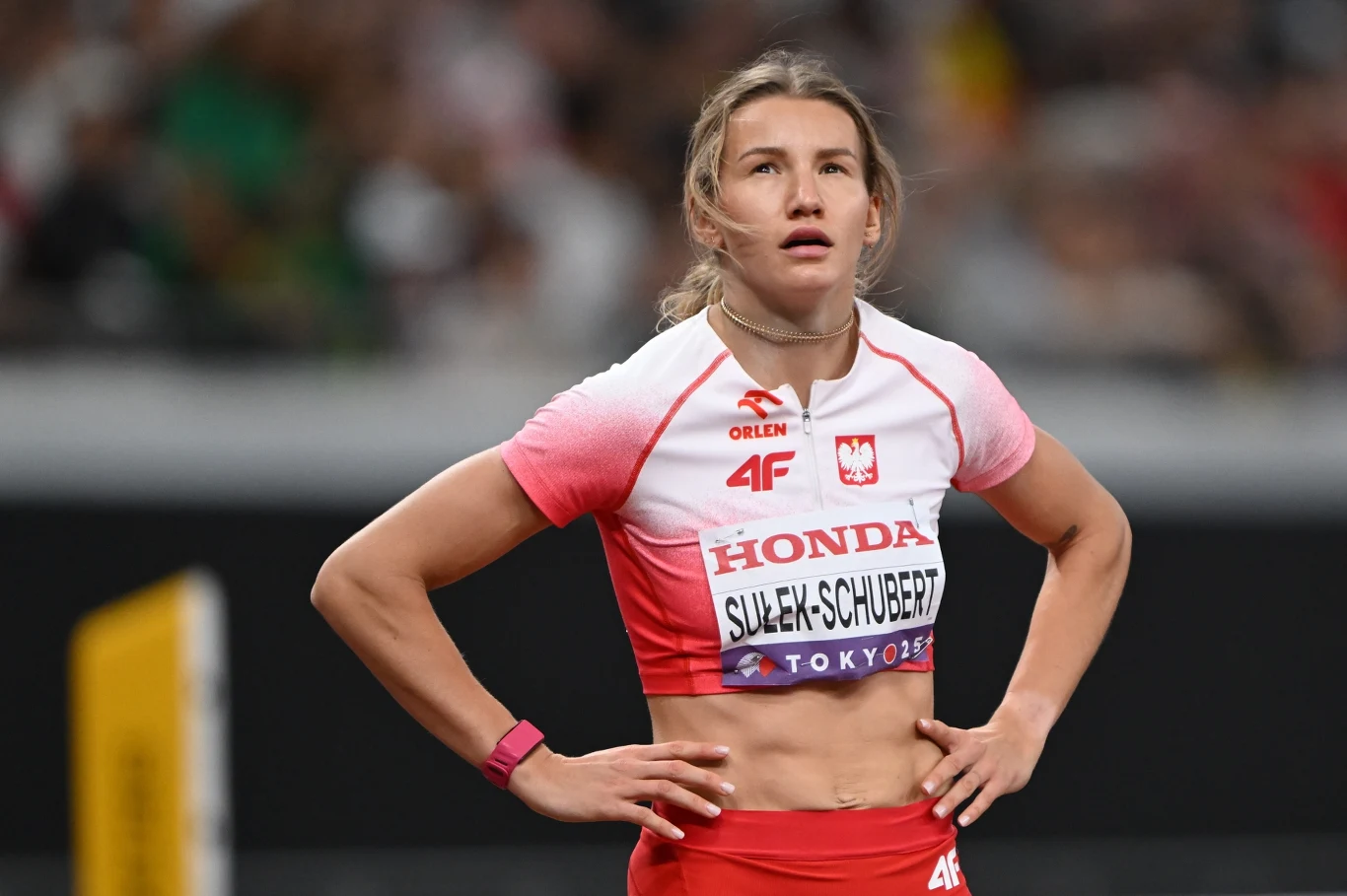 Lekkoatletka w stroju sportowym, z nazwiskiem na koszulce, stoi na stadionie, w tle częściowo rozmazana publiczność oraz elementy areny lekkoatletycznej.