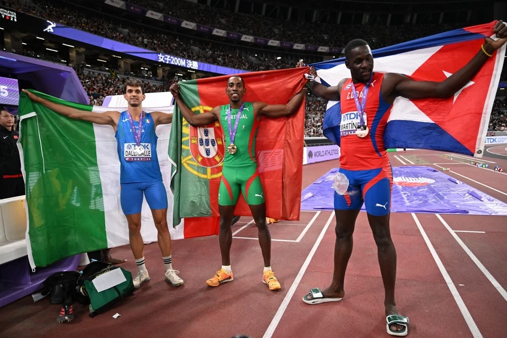 Oto najlepsi zawodnicy w konkursie trójskoku Trzech lekkoatletów stojących na stadionie lekkoatletycznym, każdy z nich trzyma flagę swojego kraju i ma medal na szyi, wokół nich znajduje się tłum widzów na trybunach.