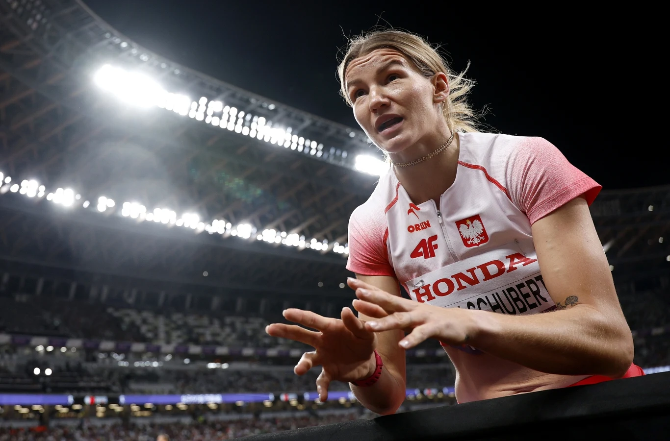 Sportsmenka w polskim stroju narodowym na stadionie lekkoatletycznym gestykuluje podczas zawodów, w tle widoczne trybuny oraz silne oświetlenie stadionowe.