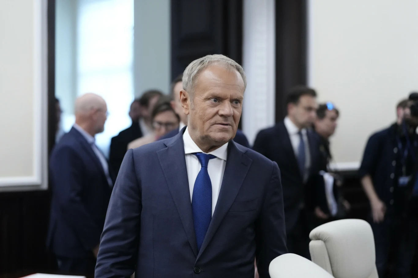 Premier Donald Tusk opublikował wideo, gdzie opiera się na niemieckojęzycznej prasie Mężczyzna w garniturze i niebieskim krawacie stoi w sali konferencyjnej, za nim widać inne osoby w formalnych strojach rozmawiające w tle.