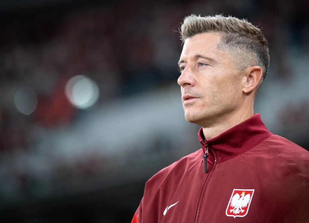 Mężczyzna w czerwonej kurtce sportowej z orłem w koronie na piersi, tło nieostre, stadion.