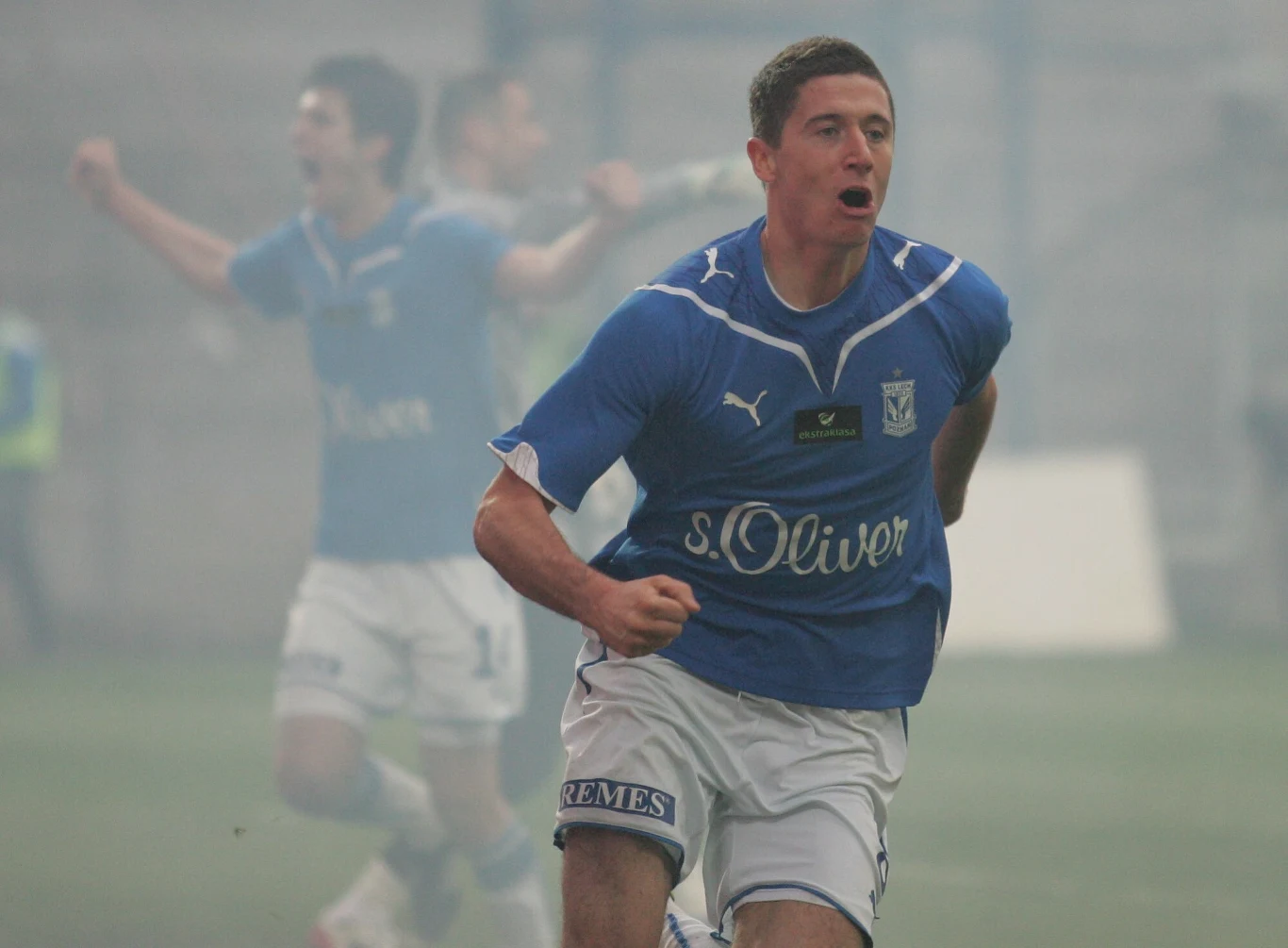 Piłkarz drużyny Lech Poznań w niebieskim stroju biegnie po boisku z emocjonalnym wyrazem twarzy, w tle drugi gracz w geście radości, atmosfera pełna dynamicznej energii sportowej.