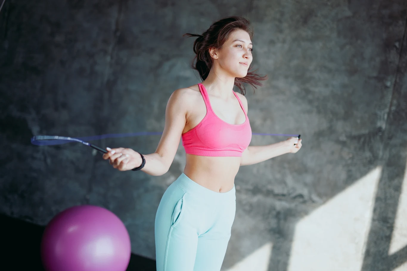 Młoda kobieta w sportowym stroju skacze na skakance w jasnym, minimalistycznym wnętrzu, obok znajduje się piłka gimnastyczna, a w tle widoczna jest szara betonowa ściana.