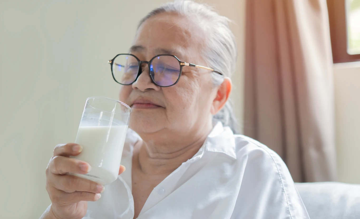 Starsza kobieta z siwymi włosami i okularami trzyma w ręce szklankę mleka, sprawiając wrażenie relaksu i zadowolenia. Ubrana w białą koszulę siedzi w jasnym pomieszczeniu z zasłoniętym oknem w tle.