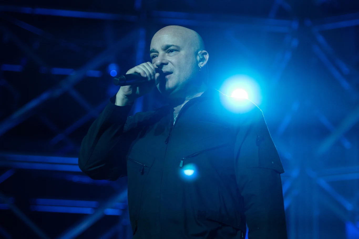 David Draiman, lider Disturbed Mężczyzna z ogoloną głową trzymający mikrofon podczas występu scenicznego, oświetlony niebieskim światłem, w tle widoczna metalowa konstrukcja sceniczna.