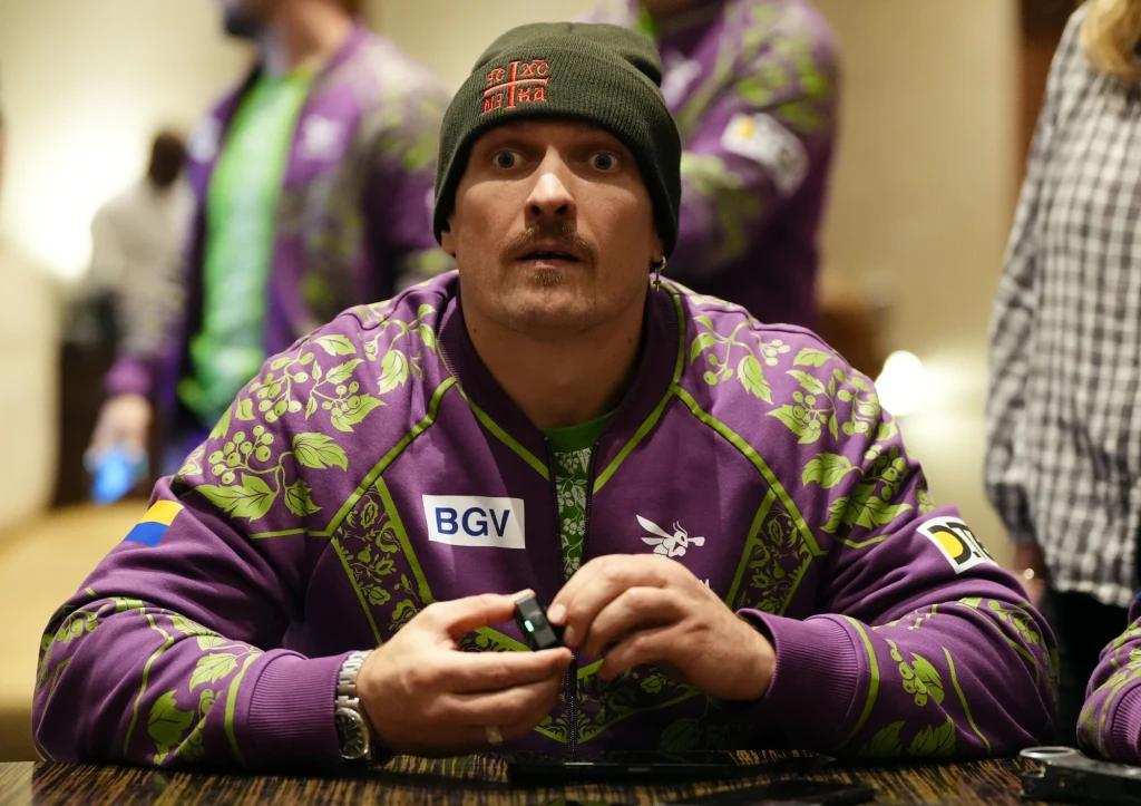 Ołeksandr Usyk Mężczyzna w zielonej czapce oraz fioletowej kurtce sportowej z zielonym motywem roślinnym siedzi przy stole i trzyma w rękach mały przedmiot, patrząc zaskoczonym wzrokiem bezpośrednio przed siebie, w tle rozmyte sylwetki innych osób.