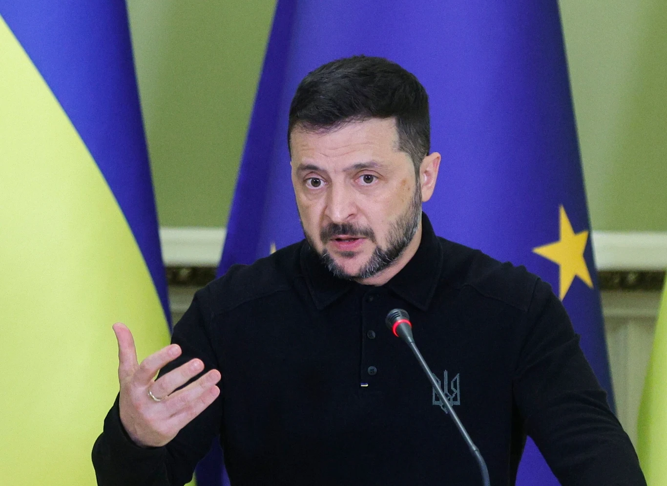 Prezydent Ukrainy Wołodymyr Zełenski Mężczyzna z krótką brodą przemawia przy mikrofonie, w tle znajdują się flagi Ukrainy oraz Unii Europejskiej.