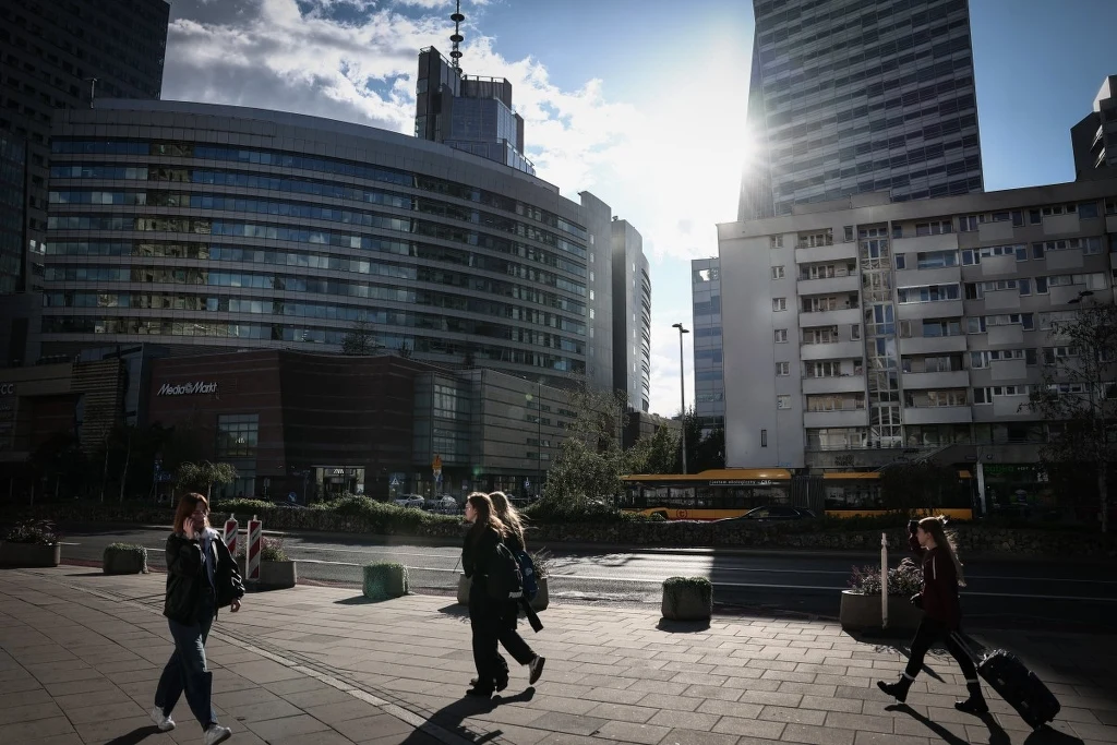 Niemieckie media: Polska goni potęgi UE (Zdj. ilustracyjne) Nowoczesne wieżowce o szklanej fasadzie w centrum miasta, chodnik z kilkoma spacerującymi osobami, jedna z nich ciągnie walizkę, w tle słońce przebijające się przez budynki.