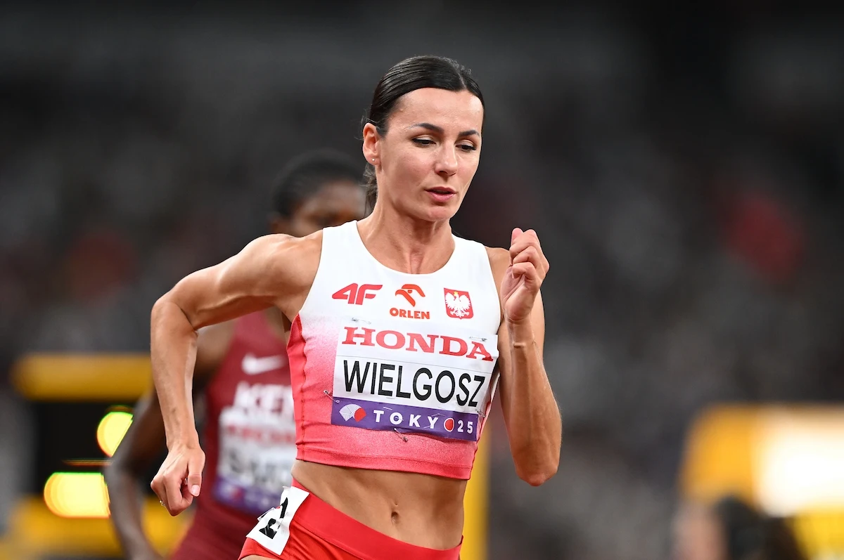 Anna Wielgosz Kobieta w biało-czerwonym stroju biegaczki z widocznym napisem na koszulce, biegnąca na stadionie lekkoatletycznym podczas zawodów sportowych, w tle widoczny inny zawodnik.