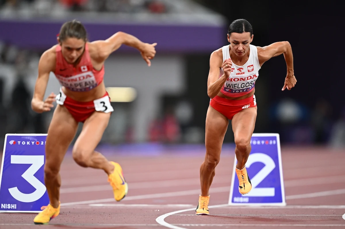 Dwie zawodniczki w strojach sportowych rozpoczynające bieg na stadionie lekkoatletycznym, intensywna koncentracja na twarzach, dynamiczne ruchy, widoczne numery na torach.
