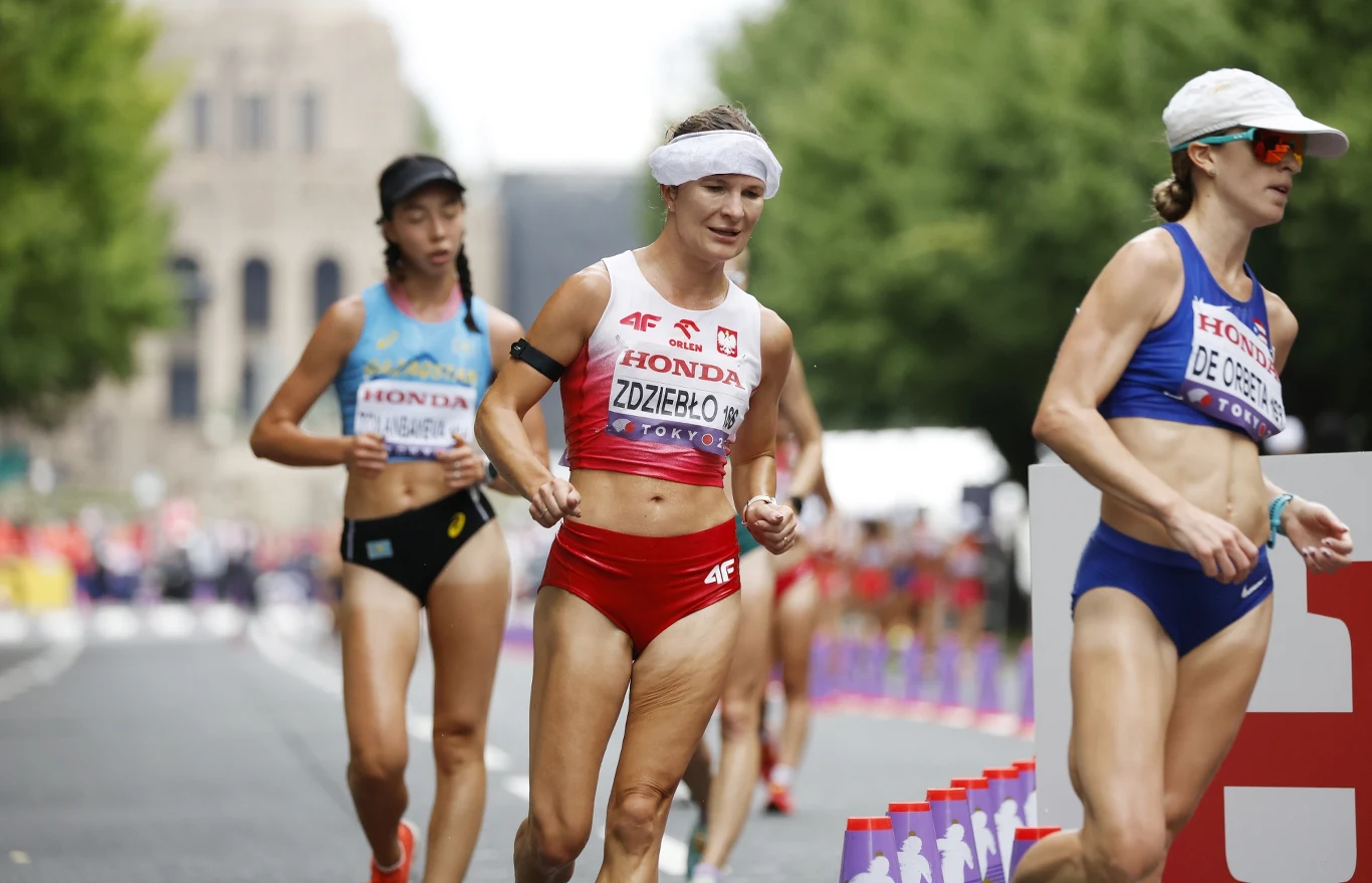 Zawodniczki biorą udział w zawodach w chodzie sportowym, ubrane w profesjonalne stroje sportowe, każda z numerem startowym oraz nazwiskiem na koszulce, jedna z nich idzie na pierwszym planie, a w tle widać innych uczestników oraz drzewa.