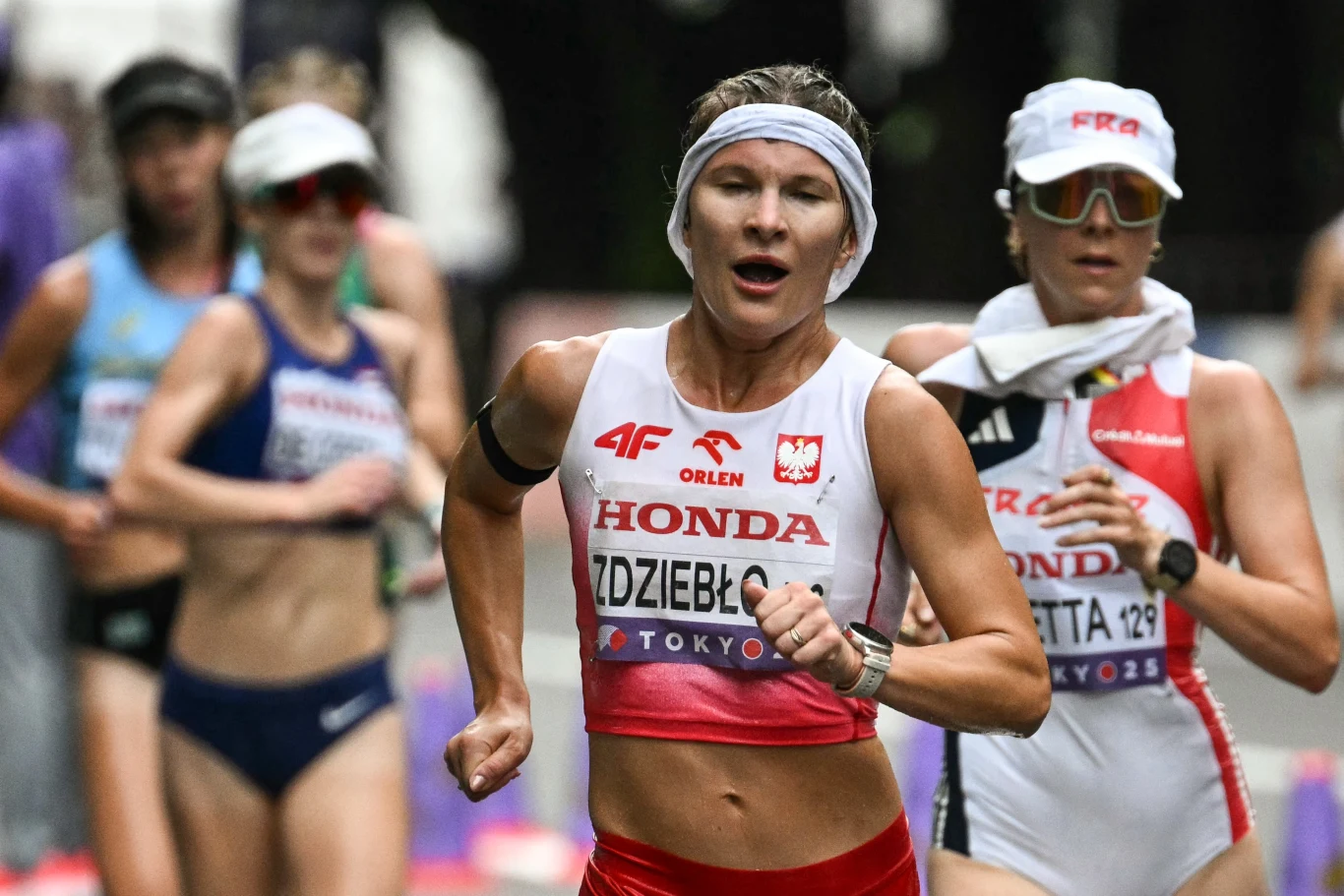 Polska lekkoatletka w stroju startowym z oznaczeniami sponsora i flagą Polski biegnie podczas zawodów, w tle inne zawodniczki uczestniczące w wyścigu chodziarskim, na sportowej trasie.