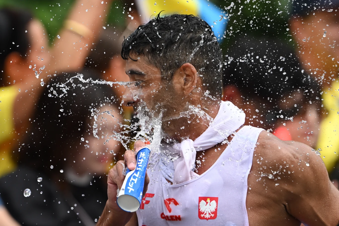 Sportowiec w białym stroju z napisem Orlen oraz godłem Polski ochładza się wodą polewając twarz podczas zawodów, w tle rozmazana publiczność.