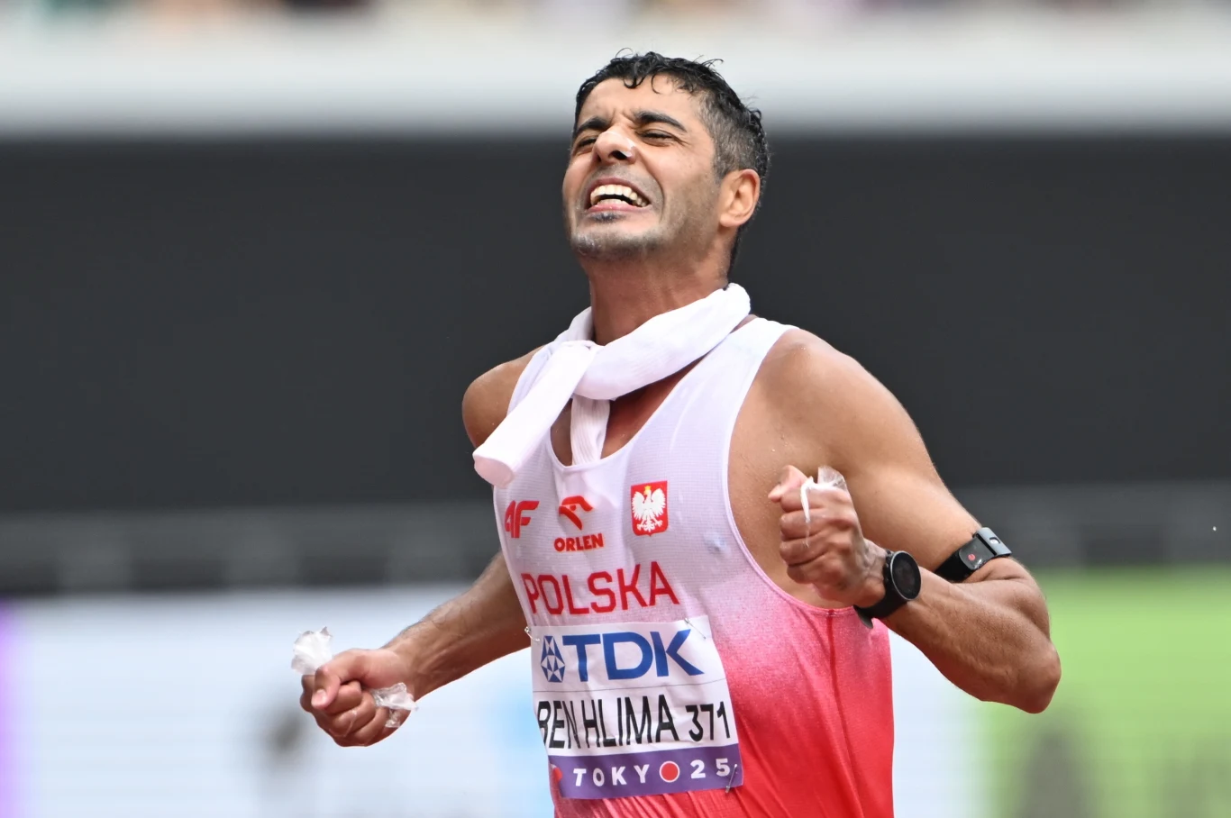 Sportowiec z Polski kończący wyścig lekkoatletyczny ze wzruszeniem i wysiłkiem widocznym na twarzy, ubrany w biało-czerwony strój z nazwą kraju i numerem startowym, owinięty ręcznikiem wokół szyi.