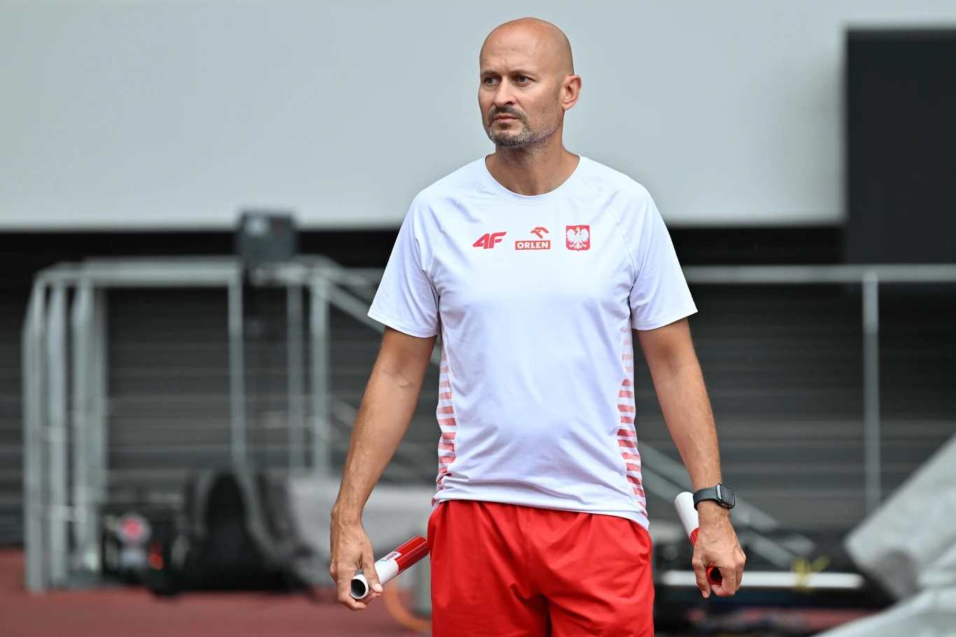 Łysy mężczyzna w sportowej koszulce reprezentacji Polski i czerwonych spodenkach, stojący na stadionie lekkoatletycznym, trzyma w dłoniach przedmioty przypominające pałeczki sztafetowe.