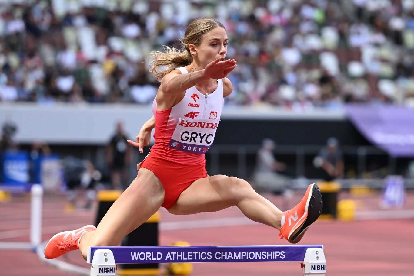 Polska lekkoatletka w czerwono-białym stroju pokonuje płotek podczas biegu przez płotki na stadionie lekkoatletycznym, w tle widoczna publiczność oraz elementy infrastruktury sportowej.