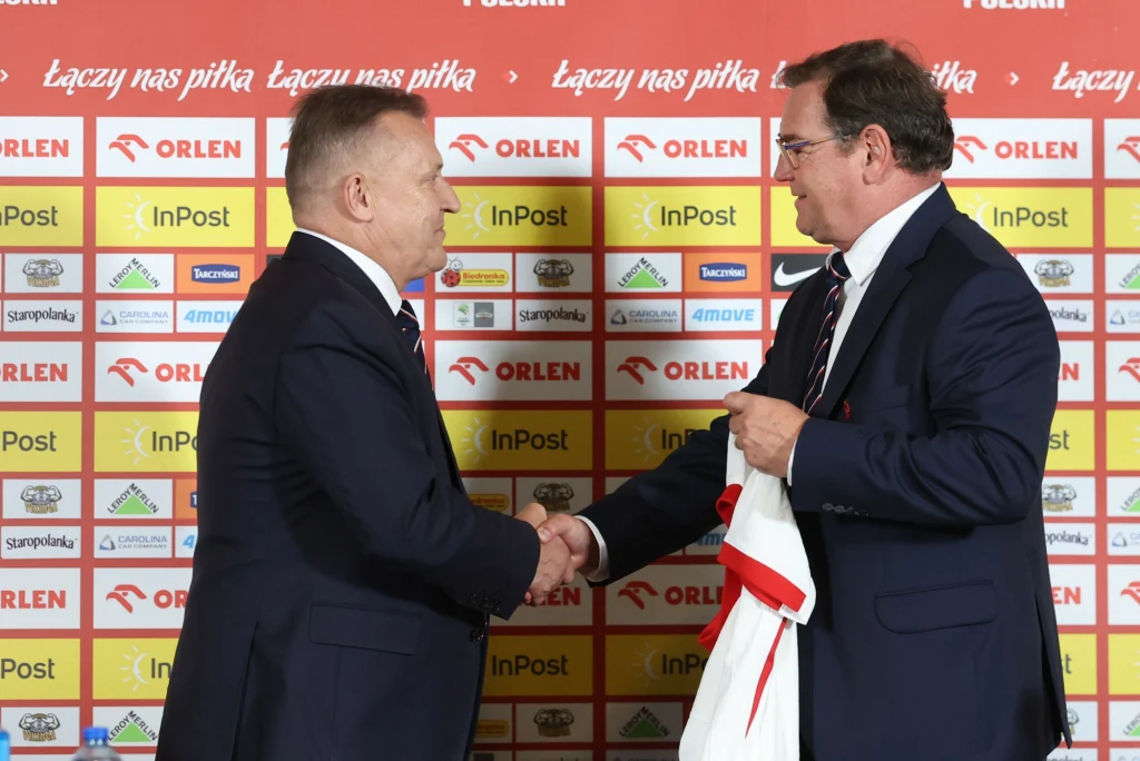 Cezary Kulesza i Jan Urban Dwóch mężczyzn w garniturach podaje sobie dłonie na tle ściany z logo sponsorów sportowych, jeden z nich trzyma biało-czerwoną koszulkę.