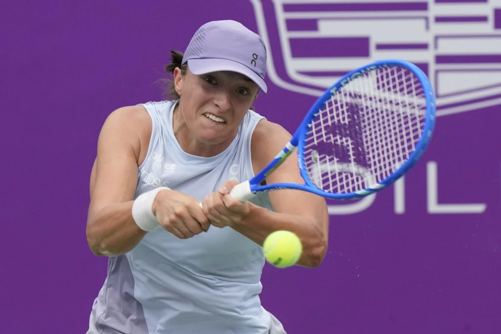 Tenisistka w ruchu uderzająca piłkę rakietą, ubrana w sportowy strój w jasnych odcieniach, na tle fioletowej ściany z logo turnieju.