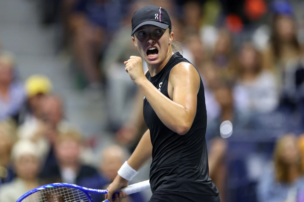 Tenisistka w czarnej odzieży sportowej podczas intensywnego momentu meczu, z wyrazem determinacji na twarzy i zaciśniętą pięścią, w tle rozmyte sylwetki widzów na trybunach.