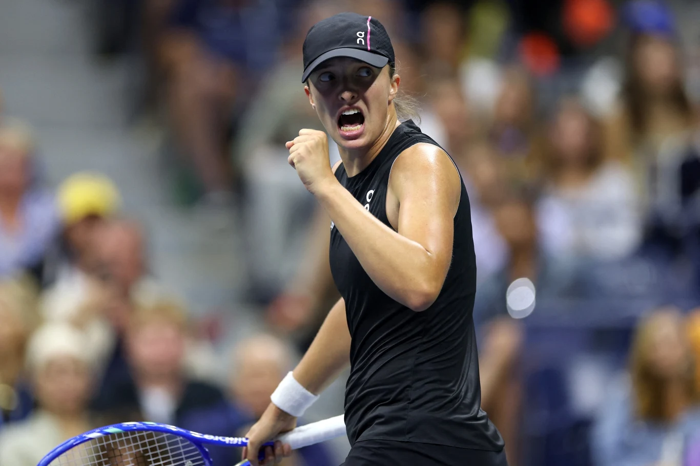 Tenisistka w czarnej odzieży sportowej podczas intensywnego momentu meczu, z wyrazem determinacji na twarzy i zaciśniętą pięścią, w tle rozmyte sylwetki widzów na trybunach.