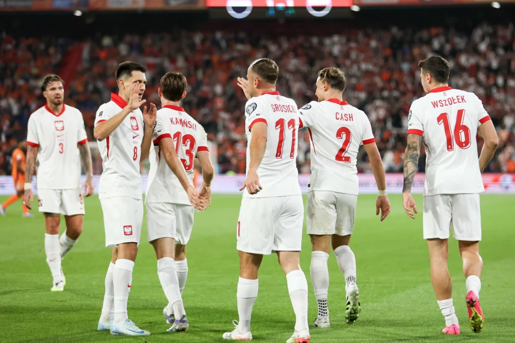 Paweł Wszołek (z prawej) Piłkarze reprezentacji Polski świętujący na boisku w trakcie meczu, ubrani w białe stroje z czerwonymi akcentami, widoczne numery i nazwiska na koszulkach, tłum kibiców w tle.