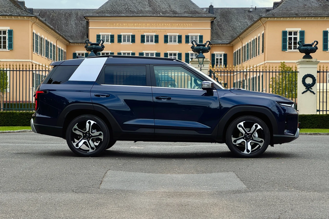 Granatowy SUV zaparkowany na tle reprezentacyjnego budynku z dużą ilością okien i symetrycznymi rzeźbami po bokach bramy.