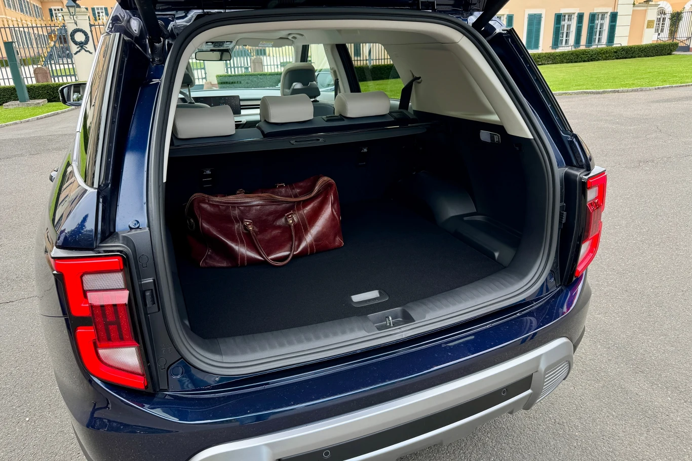 Otwarty bagażnik ciemnoniebieskiego SUV-a stoi zaparkowany na parkingu, w środku znajduje się duża bordowa torba podróżna, a w tle widać część niskiego budynku i zadbany teren zielony.