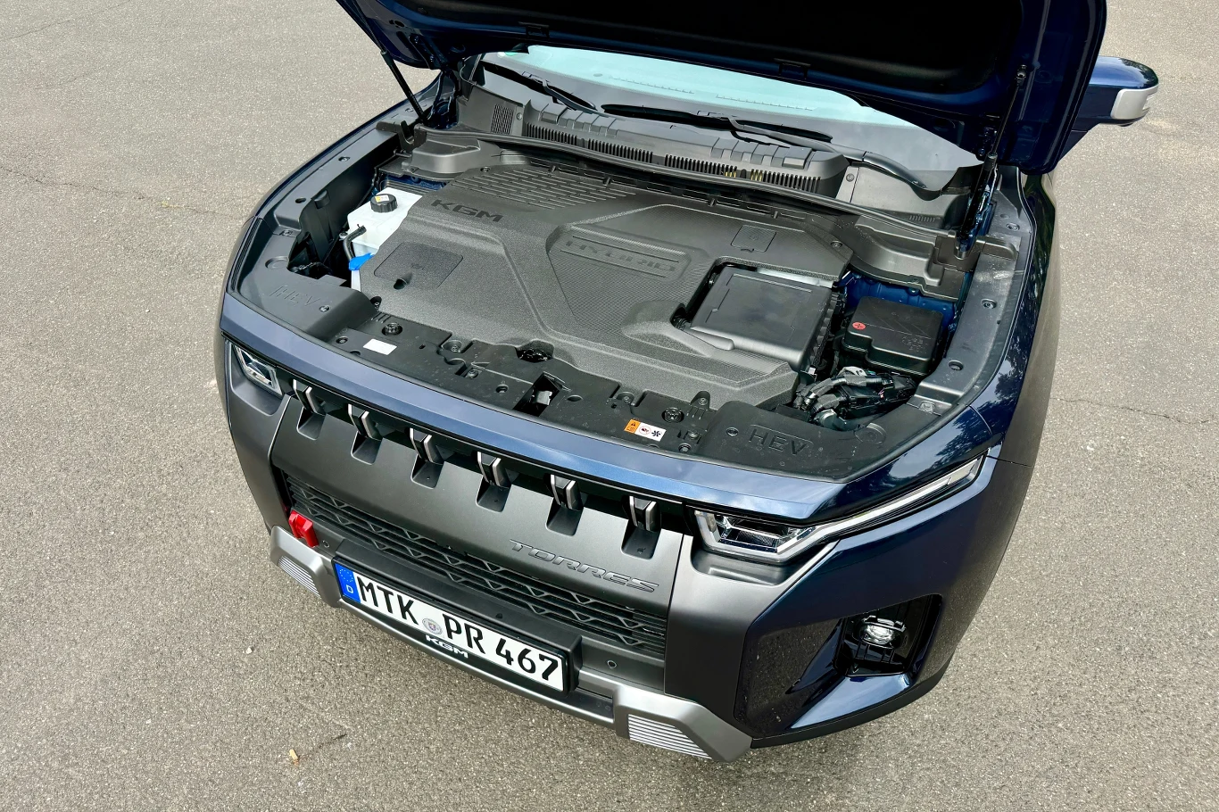 Otwarta maska samochodu terenowego w kolorze ciemnym, pod maską widoczna nowoczesna zabudowa silnika oraz elementy mechaniczne, wyraźnie widoczny przedni grill pojazdu i tablica rejestracyjna.