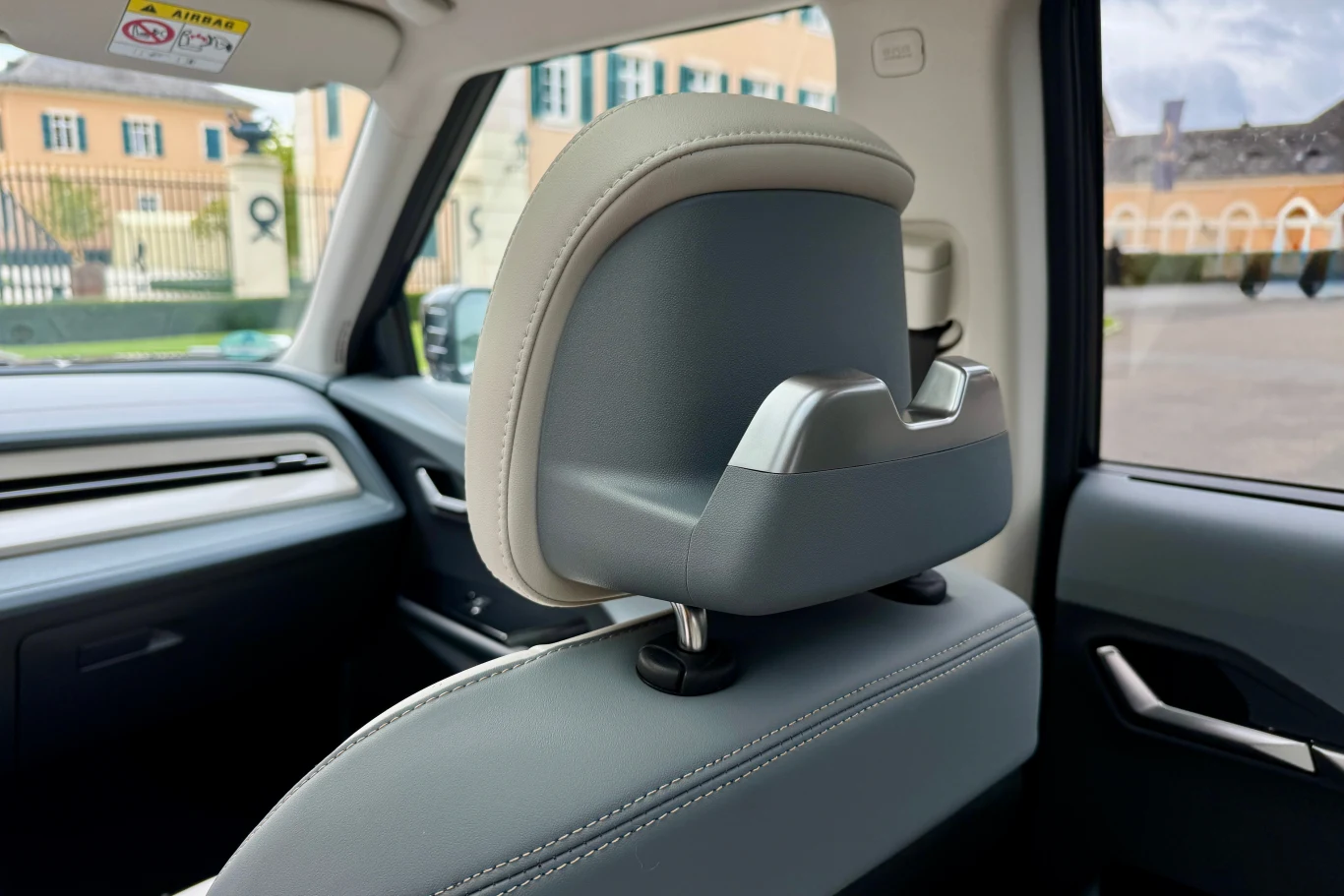 Zbliżenie na zagłówek siedzenia pasażera w nowoczesnym samochodzie, z widocznymi elementami tapicerki, fragmentem panelu drzwiowego oraz szybą, przez którą widać rozmyte budynki w tle