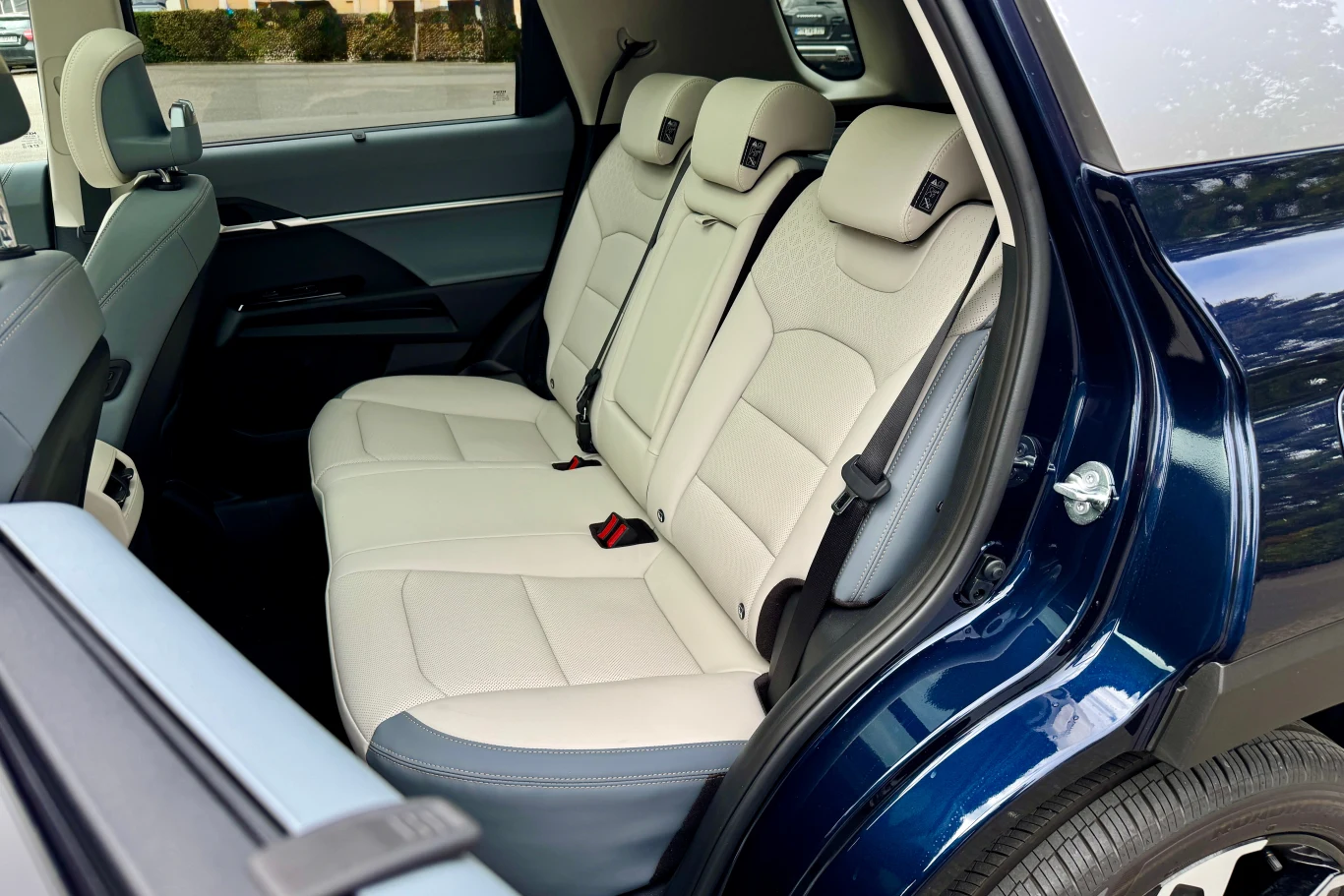 Wnętrze samochodu z otwartymi tylnymi drzwiami, jasne skórzane siedzenia w układzie trzech miejsc na tylnej kanapie, widoczne elementy pasów bezpieczeństwa oraz fragmenty karoserii pojazdu.