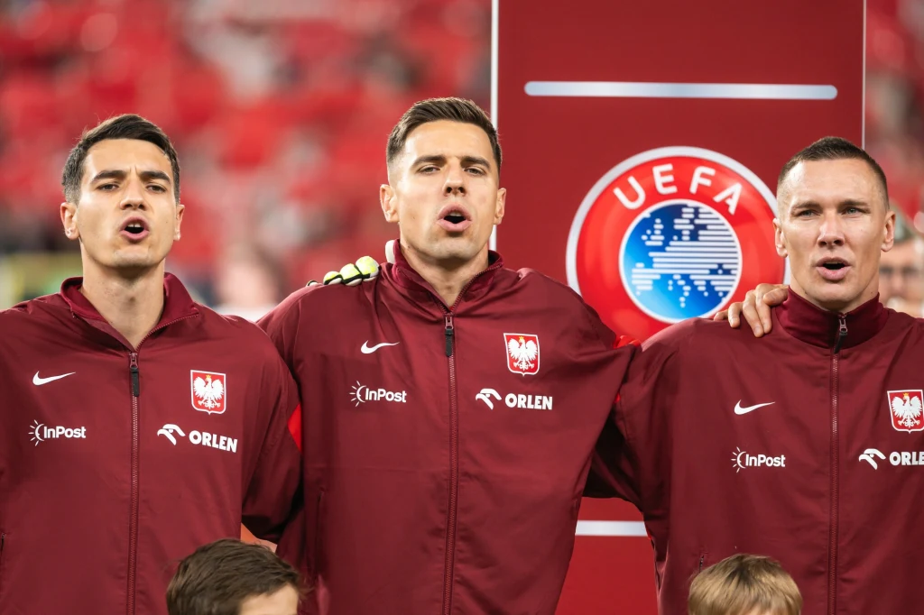 Jakub Kiwior, Jan Bednarek, Łukasz Skorupski Trzech polskich piłkarzy w czerwonych dresach reprezentacji śpiewających hymn narodowy podczas oficjalnego meczu UEFA, ręce mają położone na ramionach kolegów z drużyny, w tle widoczny jest logotyp UEFA.