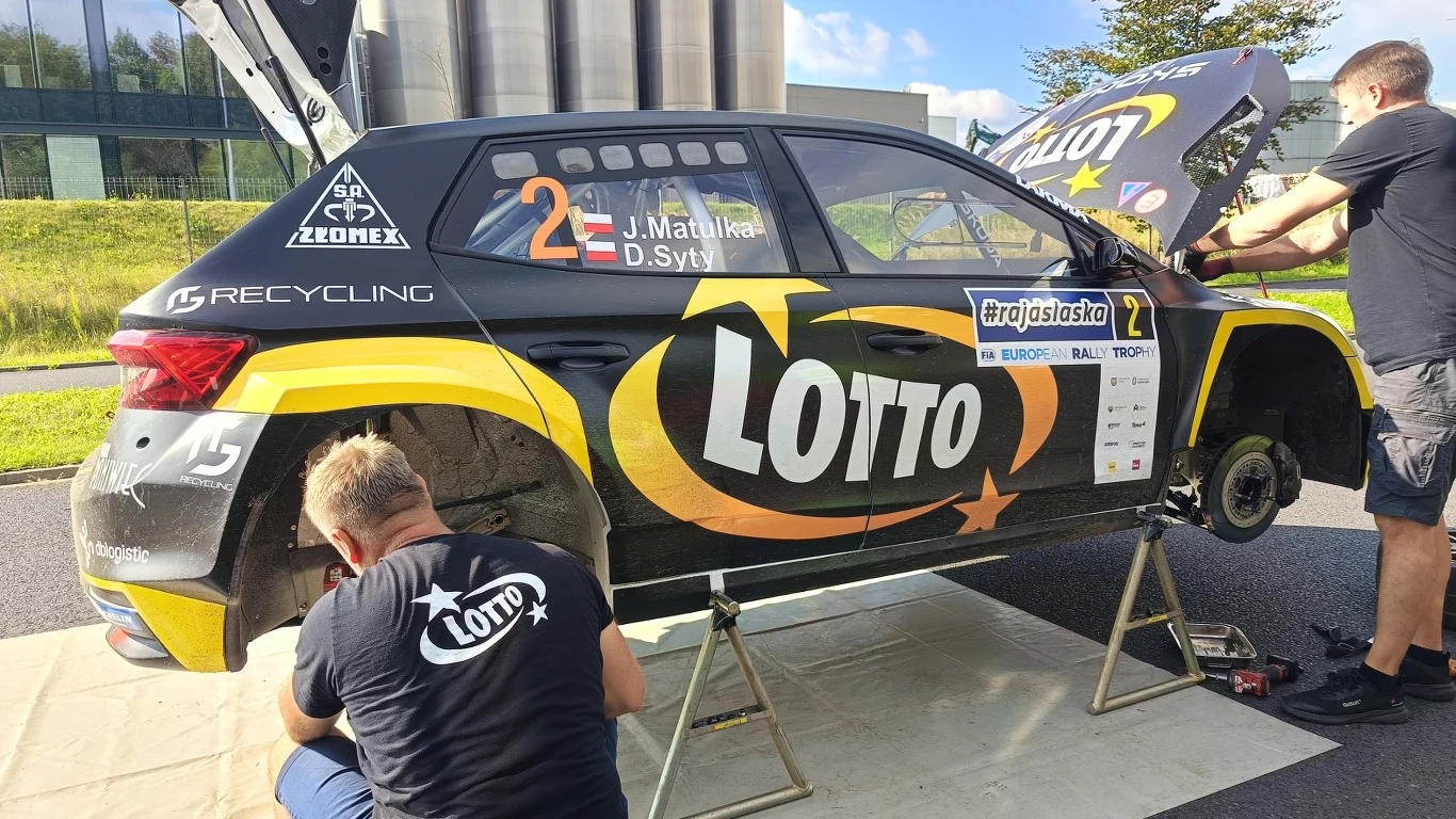 Samochód rajdowy z widocznymi logotypami sponsorów, w tym Lotto, stoi na podnośniku podczas prac serwisowych wykonywanych przez dwóch mechaników ubranych w koszulki z logo Lotto, części karoserii są otwarte, wyeksponowane są koła i silnik.