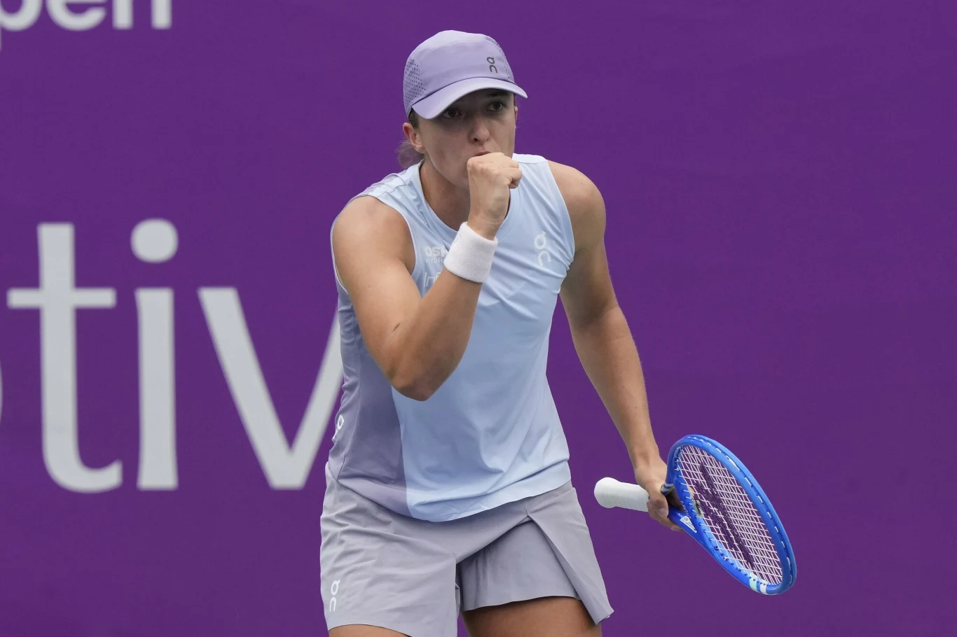 Zawodniczka tenisa ubrana w jasnofioletowy strój sportowy z rakietą tenisową w ręce świętuje punkt na tle fioletowej ścianki z białymi napisami.