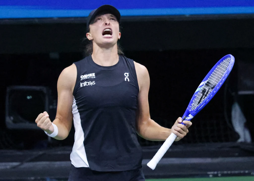 Iga Świątek Zawodniczka tenisowa ubrana w czarną koszulkę i ciemną czapkę z zaciśniętą pięścią oraz otwartymi ustami, wyrażająca silne emocje na korcie, trzymająca rakietę tenisową w lewej ręce.