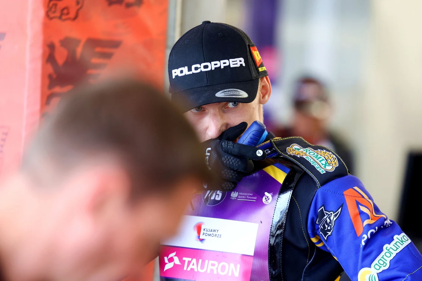 Kierowca żużlowy ubrany w kombinezon wyścigowy oraz czapkę z napisem POLCOPPER, trzymający się za część kombinezonu, z oznakami sponsorów oraz tabliczką TAURON, stojący w hali sportowej.