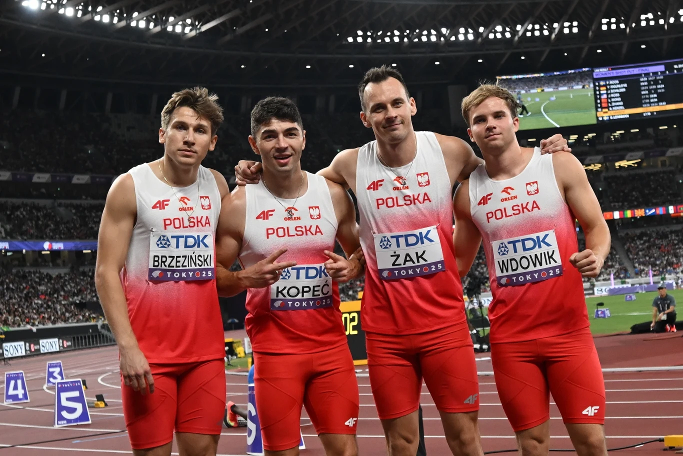 Czterech lekkoatletów w biało-czerwonych strojach reprezentujących Polskę pozuje na bieżni stadionu lekkoatletycznego, uśmiechając się do aparatu, w tle widoczna tablica wyników oraz fragmenty trybun.