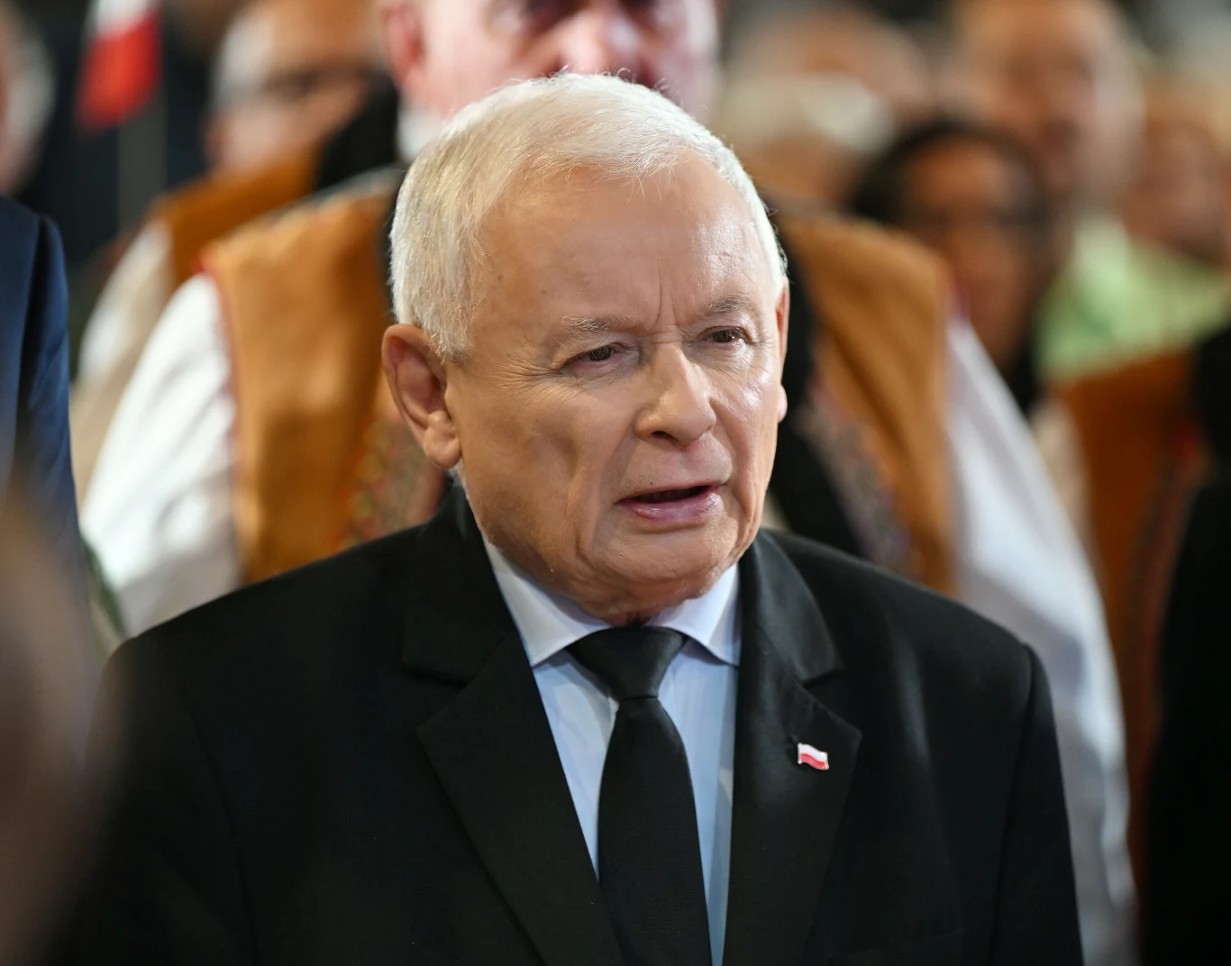 Starszy mężczyzna w garniturze z przypiętą biało-czerwoną flagą na marynarce stoi na tle grupy ludzi ubranych w tradycyjne stroje regionalne.